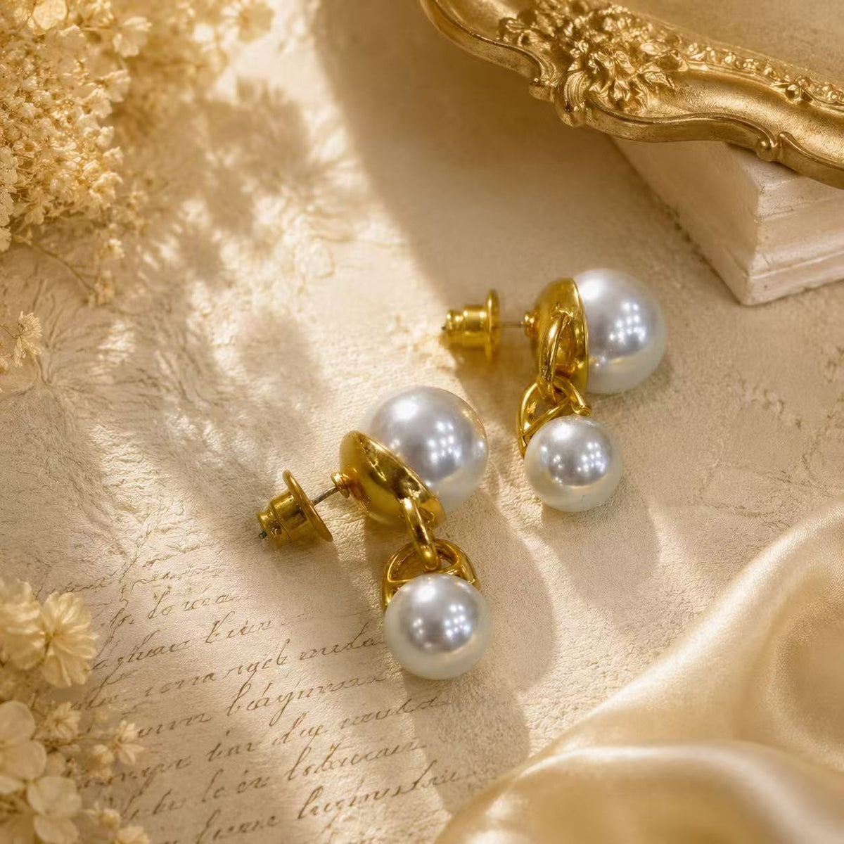 Two gold-tone pearl-style double-sphere drop stud earrings resting on cream-colored fabric