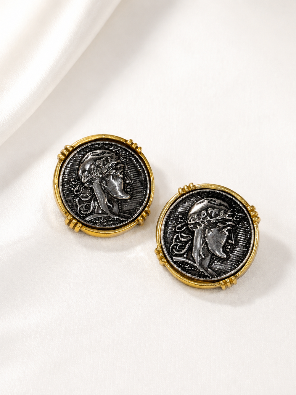Pair of gold-tone coin-inspired portrait stud earrings with dark silver-tone centers on white satin