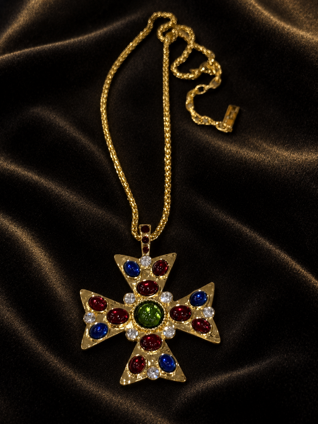 Gold-tone cross pendant necklace with red, blue, green and clear decorative stones on dark fabric.