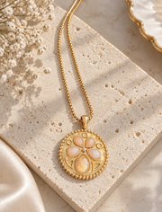 Close-up of the round gold-plated medallion pendant with peach-pink floral accents and beaded edging on a light stone display