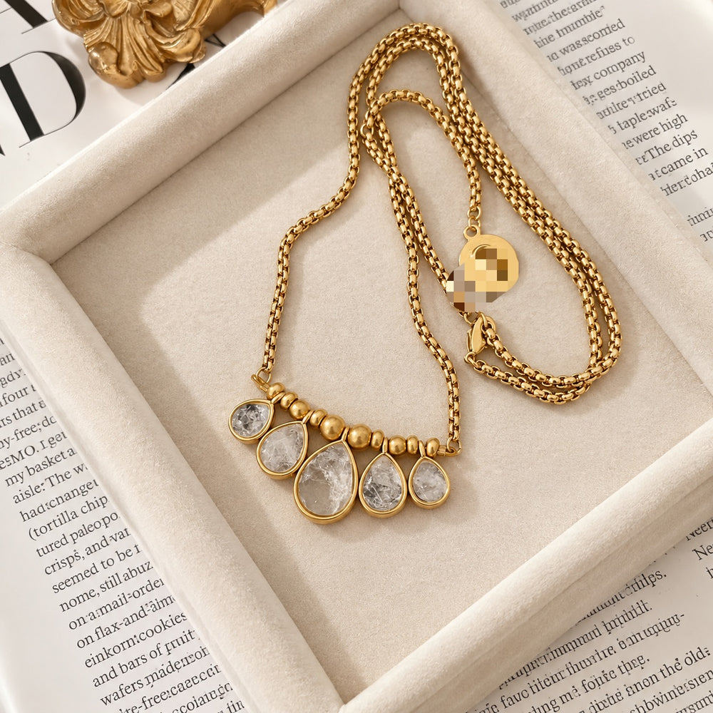 24K gold-plated teardrop crystal pendant necklace with five graduated clear pendants and gold-coloured bead accents on a cream jewellery tray