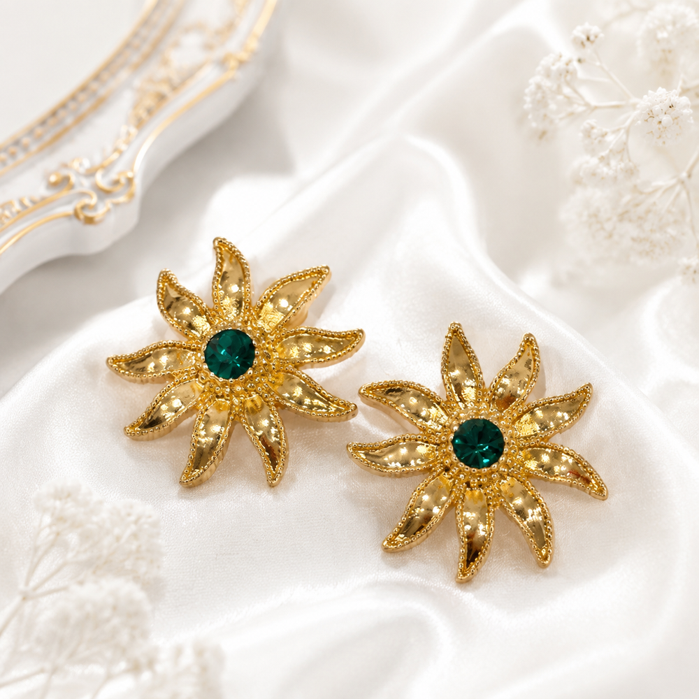 Pair of gold-tone sunburst stud earrings with deep green crystal-style centers on ivory satin