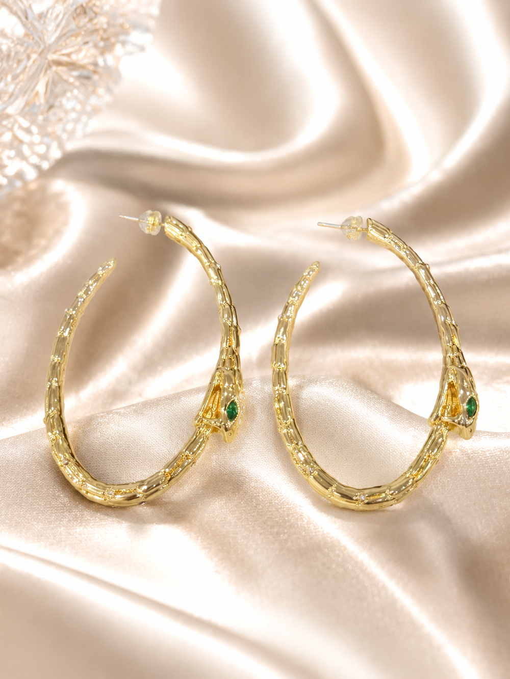 Gold-tone snake hoop earrings with green eye accents on satin background