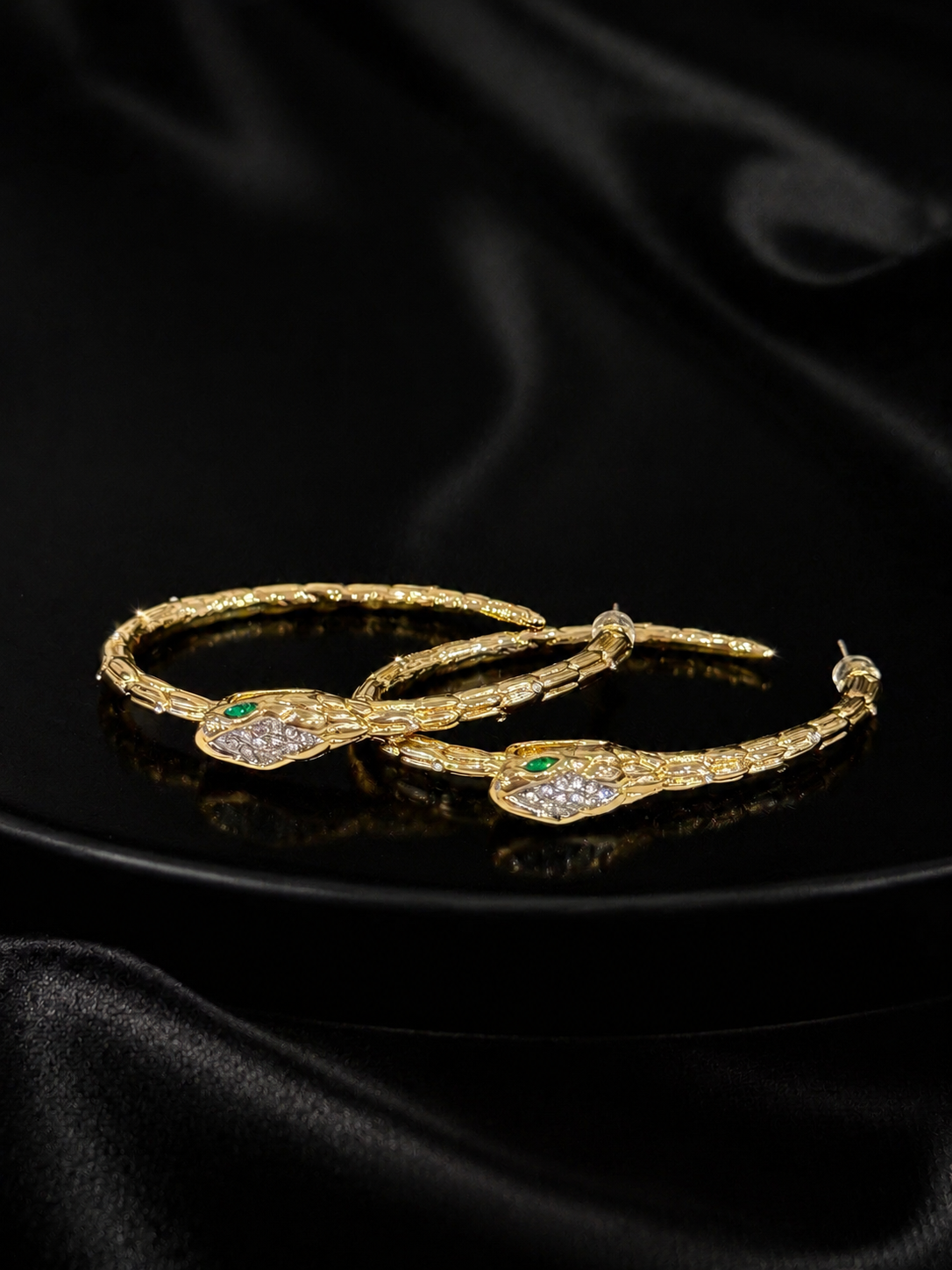 Gold-tone snake hoop earrings with green and crystal-style eye accents on black background